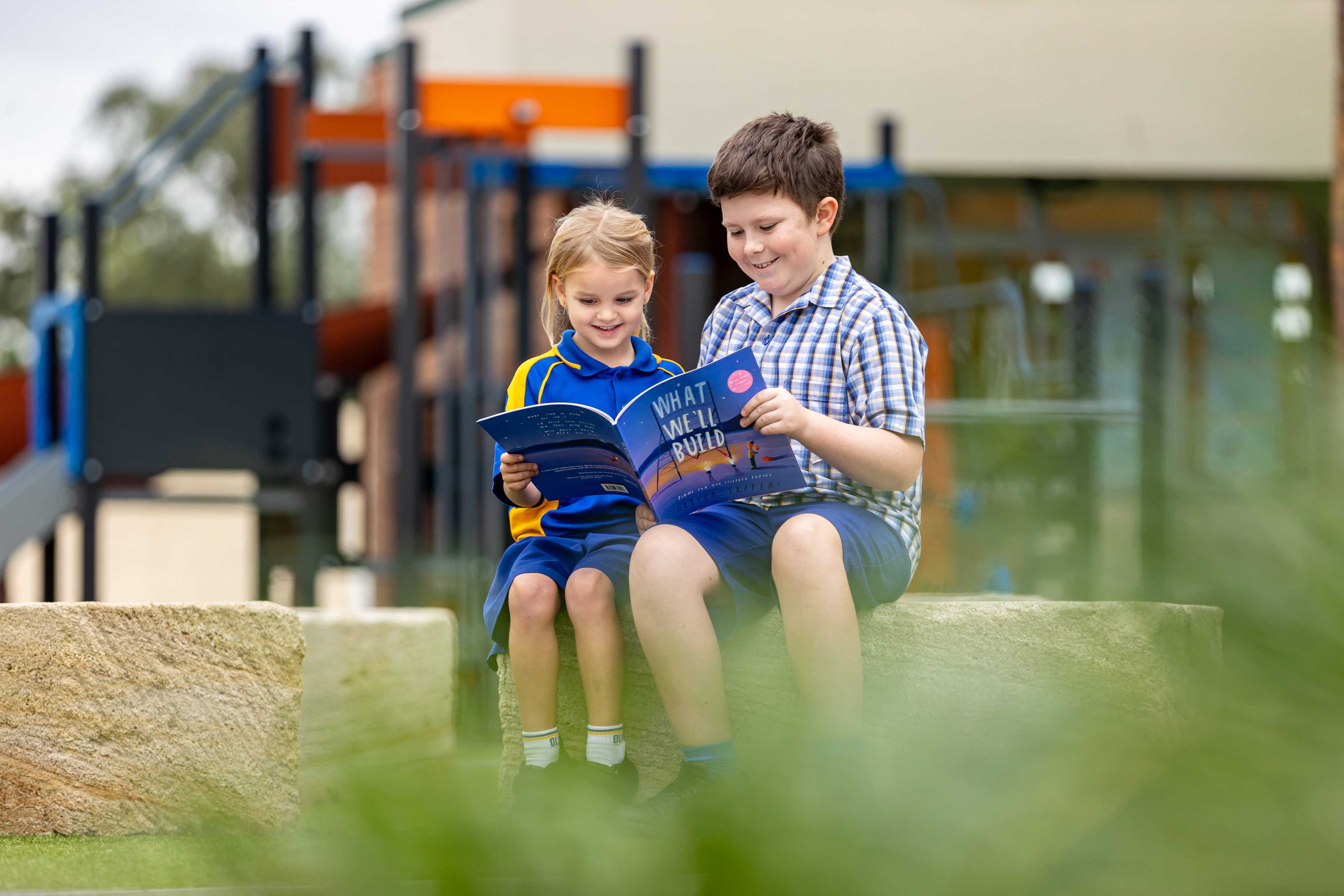Two children sitting together outdoors reading a book near a playground